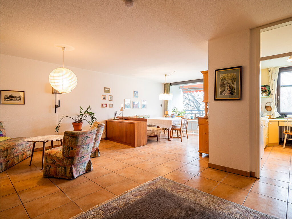 Ein geräumiges Wohn- und Esszimmer mit Terrakotta-Fliesen, gemusterten Polstermöbeln, einer großen runden Papierlampe und einem Holz-Sideboard. Im Hintergrund befindet sich ein Essbereich am Fenster, rechts ist ein offener Durchgang zu einer Küche.